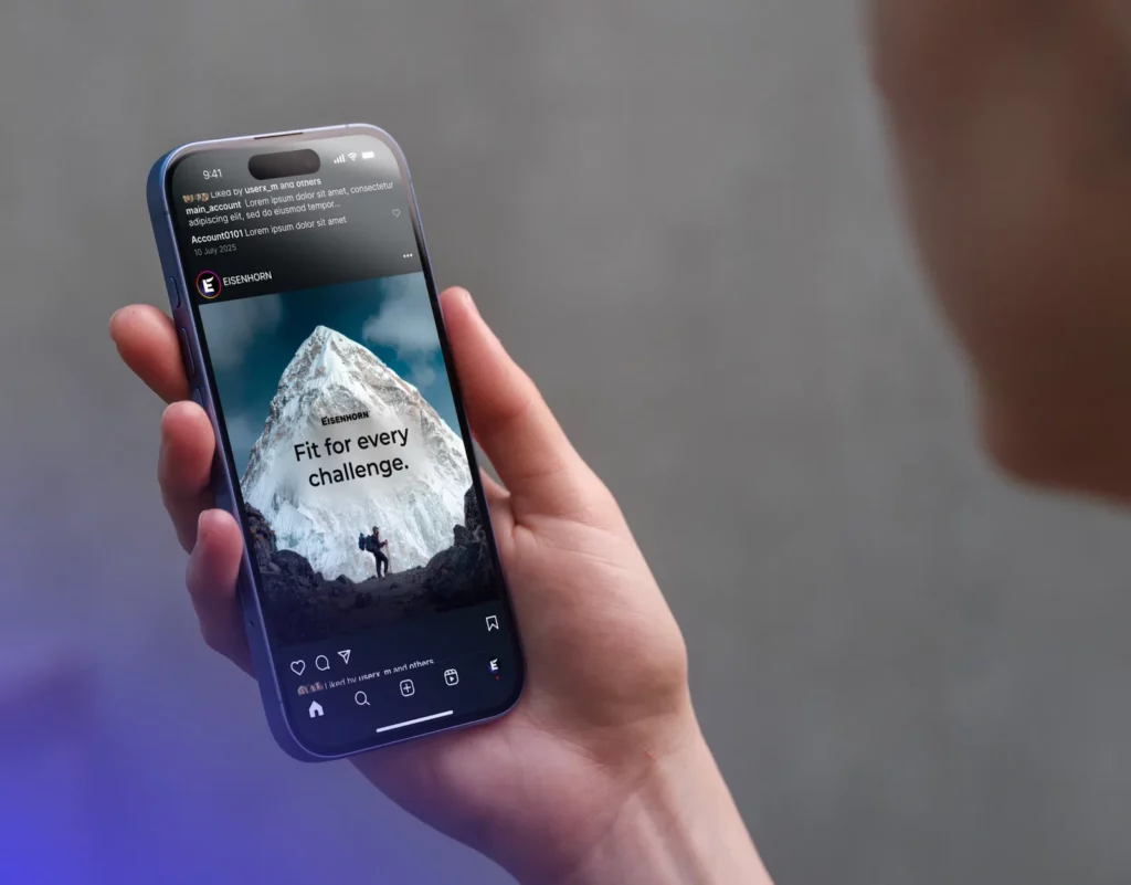 A person holds a smartphone displaying an Instagram ad for EISENHORN. The ad shows a mountain climber scaling a snowy peak with the text "Fit for every challenge." The post includes engagement details and appears to be well-liked.