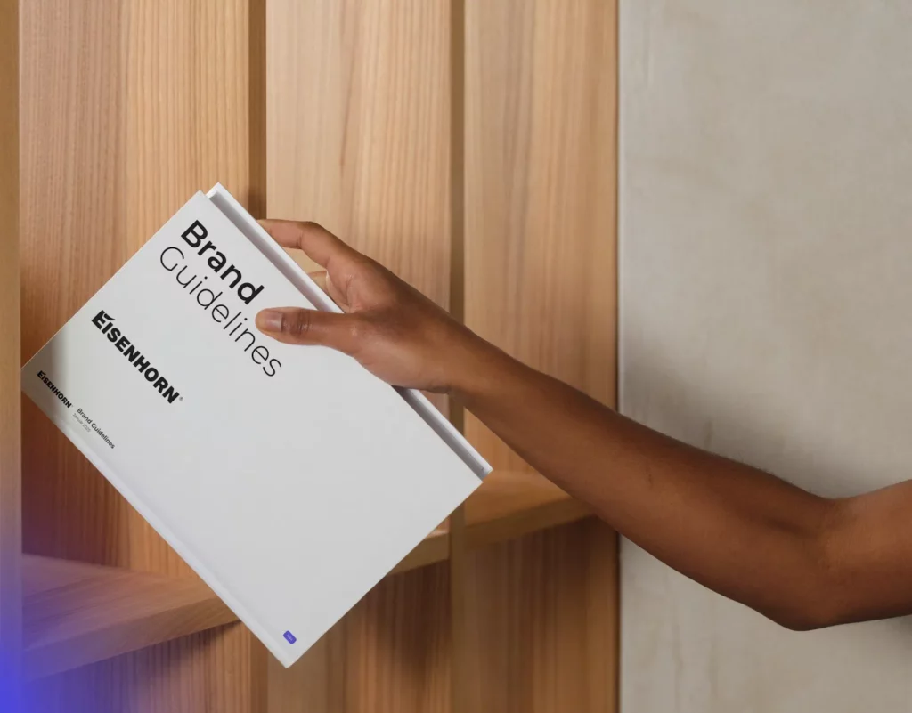 A person is placing a book titled "Brand Guidelines" by EISENHORN on a wooden shelf. The book is white with black text, emphasizing branding standards.