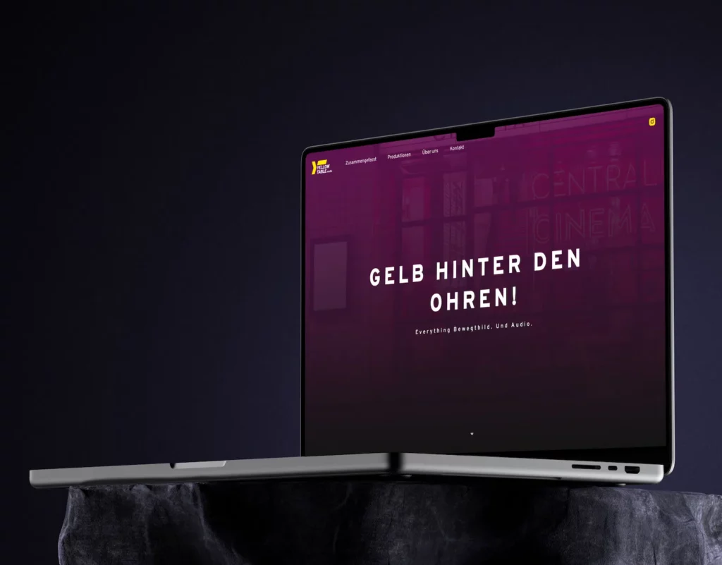 A sleek laptop displaying the Yellow Table Media website design. The screen features a purple background with bold white text, "GELB HINTER DEN OHREN!" and "Everything Bewegtbild. Und Audio." Menu options are visible at the top.